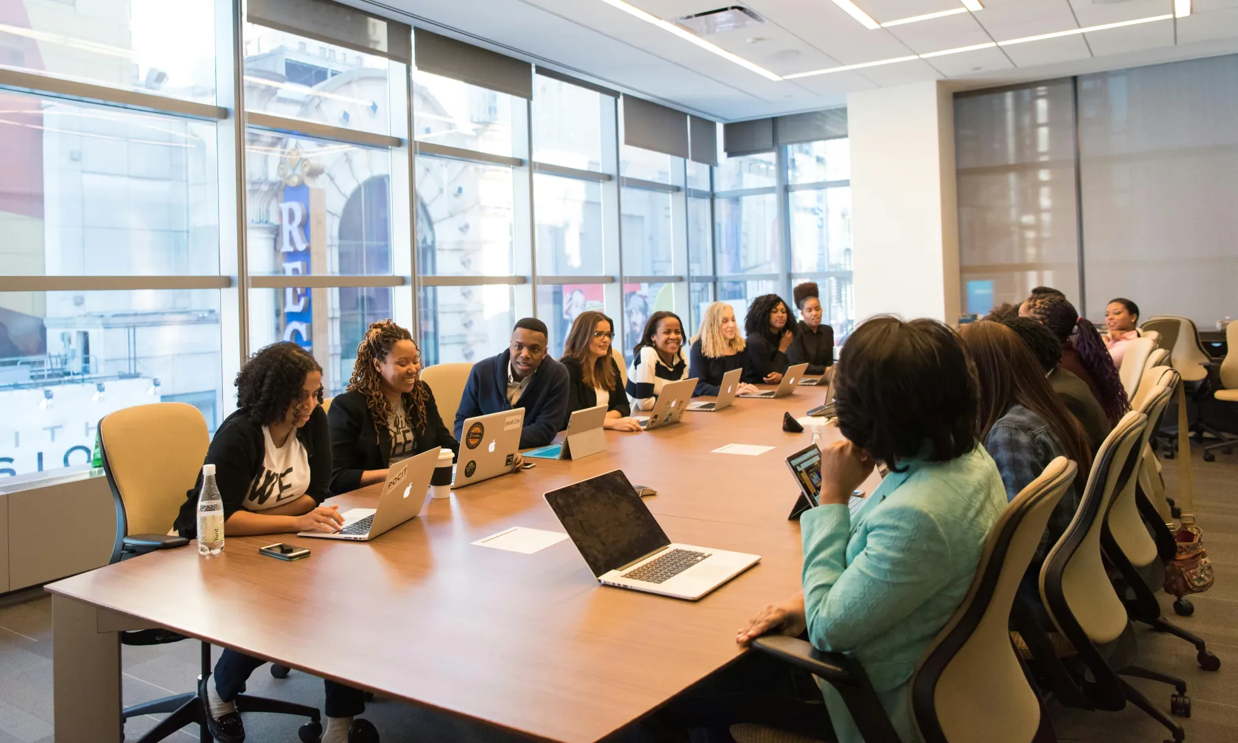 A diverse group of people in a modern conference room, sitting around a large table with laptops, engaged in a meeting